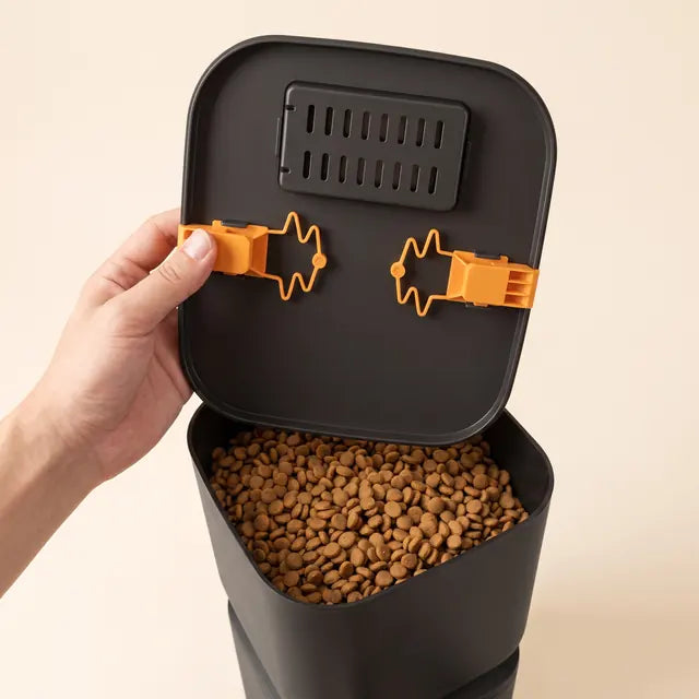 A hand opening the lid, showing the two orange locking clips and the desiccant box inside the lid, with kibble in the container.
